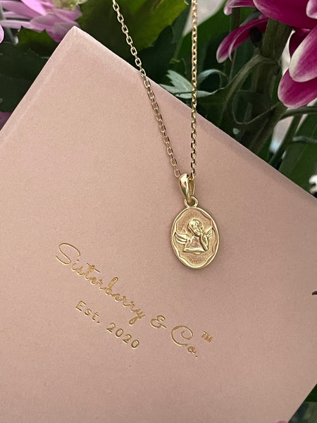 A gold pendant necklace with an oval guardian angel medallion, displayed on a pink presentation gift box from 'Sisterberry & Co' with pink flowers in the background.