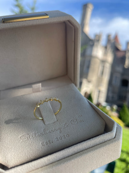 A delicate gold ring with small clear stones set on a band, displayed inside an open beige jewelry box. The box is embossed with the brand name 'Sisterberry & Co.