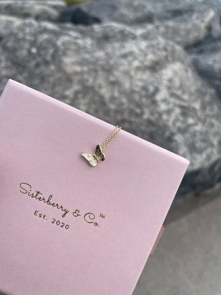 A gold butterfly pendant necklace rests on a light pink Sisterberry & Co. gift box, set against a blurred gray rock background.