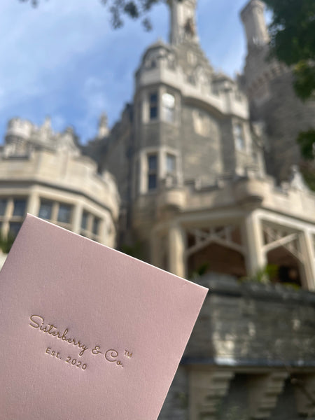 A close-up of a dusty pink textured card or envelope embossed with the brand name 'Sisterberry & Co Est. 2020', held in the foreground with a blurred castle-like building and blue sky in the background.