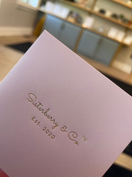 A close-up of a light pink textured card or envelope with gold foil text reading 'Sisterberry & Co. Est. 2020'