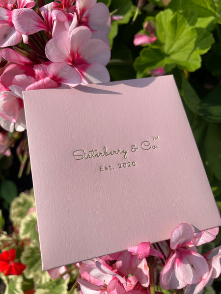 A square light pink card with the brand name 'Sisterberry & Co.' and 'Est.2020' on a bed of pink flowers outside in a garden