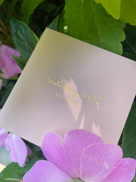 A square pale pink card or packaging piece with gold foil text reading 'Sisterberry & Co Est. 2020', photographed among green leaves and pink flowers.