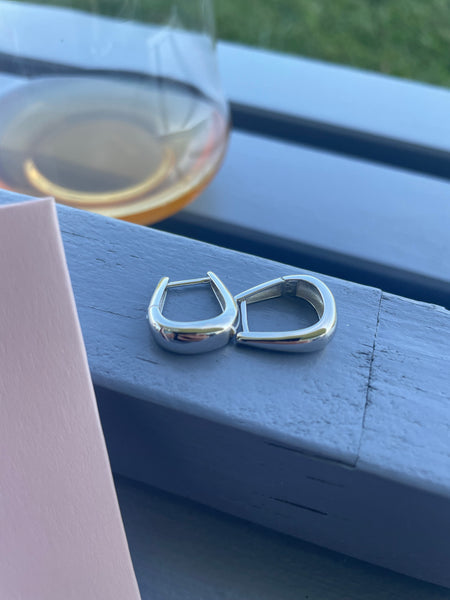 A pair of polished silver hoop earrings resting on a blue-purplish wooden bench, with a glass of amber wine blurred in the background and a pink card edge visible on the left.