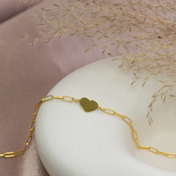 A delicate gold paperclip chain bracelet with a small polished gold heart charm, displayed on a white ceramic bust against a soft pink satin background with dried grass stems.