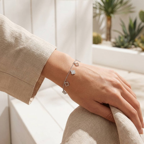 A delicate silver charm bracelet with three small heart-shaped charms worn on a person's wrist, shown in a bright, modern indoor setting with a beige linen outfit.