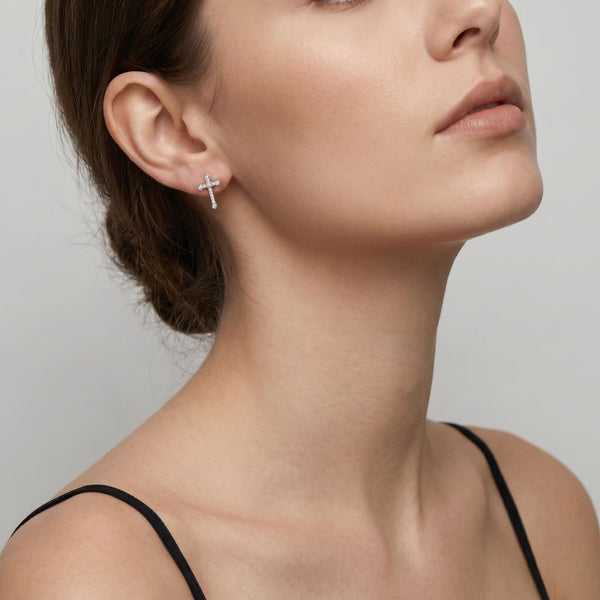 A close-up of a model's ear and neck wearing a small silver cross stud earring against a light gray background.